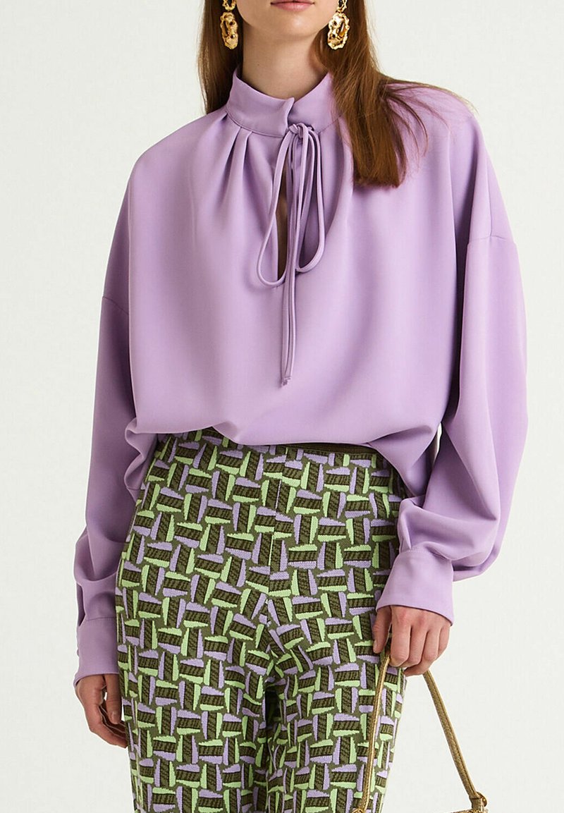 Blusa color lavanda a collo alto con dettaglio a fiocco, abbinata a pantaloni verde e viola con motivo geometrico.