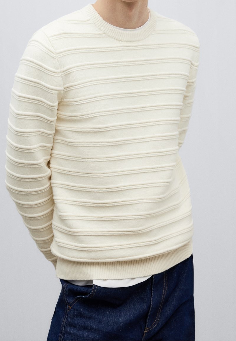 Personne portant un pull côtelé couleur crème avec des rayures horizontales texturées et un jean bleu foncé sur un fond uni.