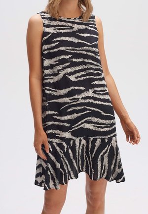 Ärmelloses schwarzes Kleid mit weißen unregelmäßigen Zebrastrifen und ausgestelltem Saum, getragen von einer stehenden Frau.