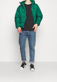 Zelená polstrovaná bunda s kapucí, černé tričko pod ní, modré džíny a černé tenisky s bílými špičkami. Texturované materiály.