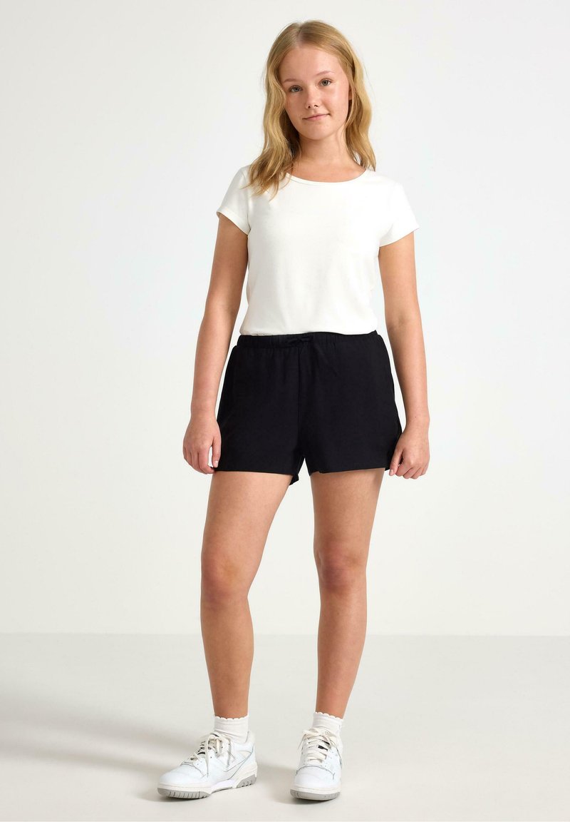 Hvid kortærmet top kombineret med sorte shorts med elastisk talje. Modellen bærer hvide sneakers og hvide ankelsokker. Simpel og afslappet outfit.