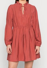 Abito rosso con texture a coste, colletto alto voluminoso, scollatura a occhio di pernice e maniche lunghe. L'elastico in vita crea una silhouette aderente.