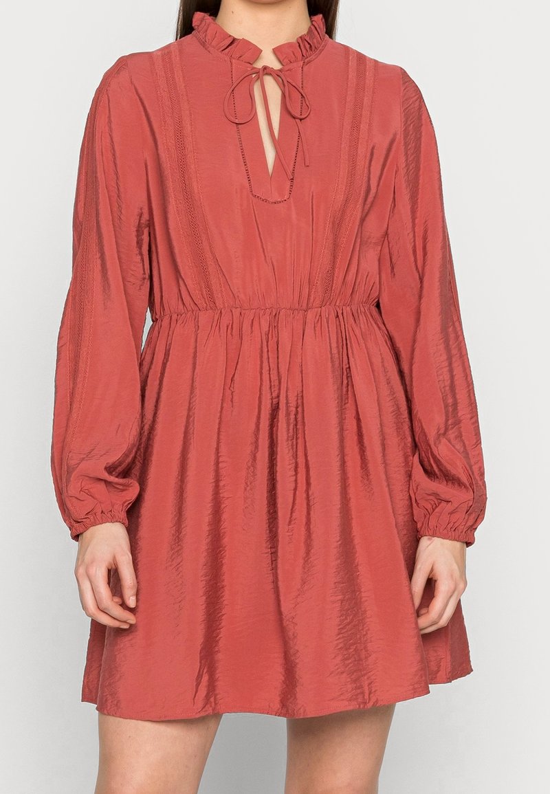 Abito rosso con texture a coste, colletto alto voluminoso, scollatura a occhio di pernice e maniche lunghe. L'elastico in vita crea una silhouette aderente.