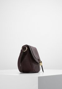 Tmavě hnědá kožená taška přes rameno s klopou, zlatými kroužkovými úchyty a malým kulatým zlatým přívěskem, vystavená na bílém podstavci.