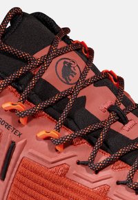 Chaussure de randonnée rouge-orange avec accents noirs, tige texturée et système de laçage distinctif. Présente un logo et une marque GORE-TEX.