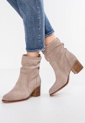 Stivaletti alla caviglia in suede beige con design morbido, caratterizzati da una cucitura decorativa e tacco in legno impilato. Indossati con jeans blu alla caviglia.