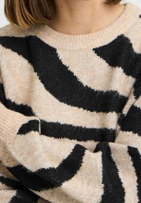 Pull beige avec de larges rayures noires ondulées. Tissu doux et texturé avec un col et des poignets côtelés. Gros plan mettant en valeur le motif et la douceur.