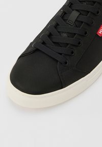 Czarny tekstylny sneaker z zaokrąglonym noskiem, płaskimi sznurowadłami i białą gumową podeszwą. Posiada małą, czerwoną metkę z logo blisko sznurowadeł.