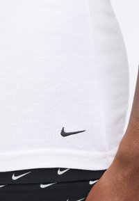 Κοντινό ενός ατόμου που φοράει άσπρο ριμπ πουκάμισο με μικρό μαύρο λογότυπο της Nike κοντά στο τελείωμα και μαύρο παντελόνι με επαναλαμβανόμενα λευκά λογότυπα της Nike.