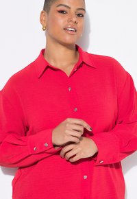 Smiling person with short hair wears a bright red button-up shirt with decorative buttons, standing against a white background.