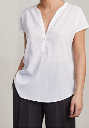 Femme portant un chemisier blanc à manches courtes avec un col en V et des boutons, associé à un pantalon noir ample, debout avec une main dans la poche.