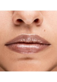 Gros plan sur des lèvres avec une teinte brun clair glossy. Texture lisse avec un léger éclat, fond au ton de peau naturel. Pas de motifs ni d'accents visibles.