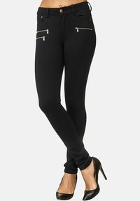 Zwarte skinny broek gemaakt van rekbaar materiaal, met twee ritssluitingen aan de voorkant en een hoge taille. Gekleed met zwarte hoge hakken.