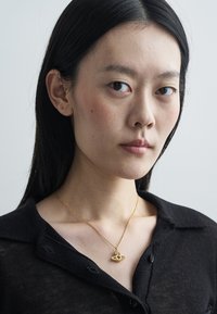 Gold necklace with intricate pendant, worn over a black button-up shirt. Smooth skin and dark hair complement the accessories.