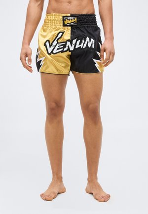 Άνδρας που φοράει μαύρα και χρυσά σορτς Venum με μοτίβα κεραυνού, όρθιος ξυπόλυτος σε απλό φόντο.