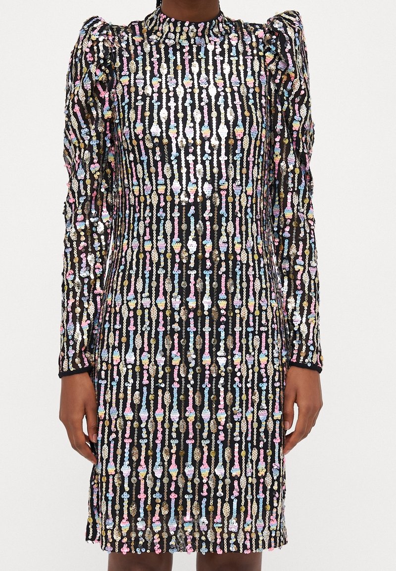 Robe noire à manches longues avec des rayures verticales de paillettes multicolores et des épaules bouffantes, portée par une personne les mains détendues sur les côtés.
