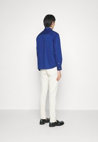 Camisa azul de manga larga con puños de botón, con corte relajado, textura suave, combinada con pantalones blancos y mocasines negros. Vista trasera.