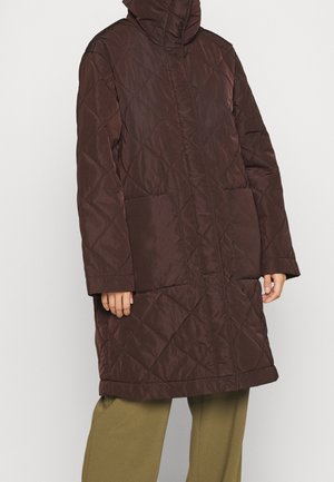 Brązowy pikowany długi płaszcz z kapturem i przednimi kieszeniami naszywanymi, noszony z oliwkowozielonymi spodniami, widoczny od ramion do połowy uda.