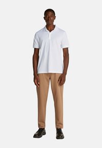 Polo bianco, maniche corte e colletto abbinato a pantaloni beige. Il modello indossa scarpe nere. Design semplice, senza motivi o logo.