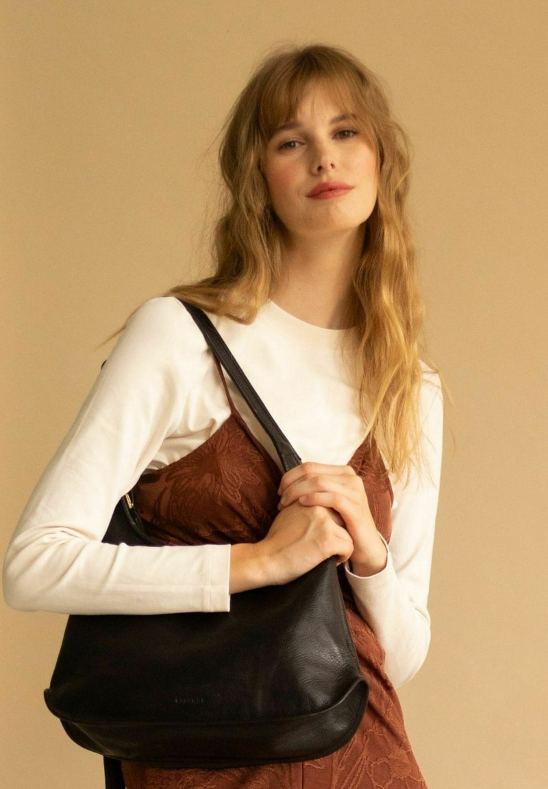 Czarna skórzana torebka o miękkiej fakturze, zakrzywionym kształcie i pojedynczym, regulowanym pasku. Modelka ma na sobie białą bluzkę z długim rękawem i brązową sukienkę.