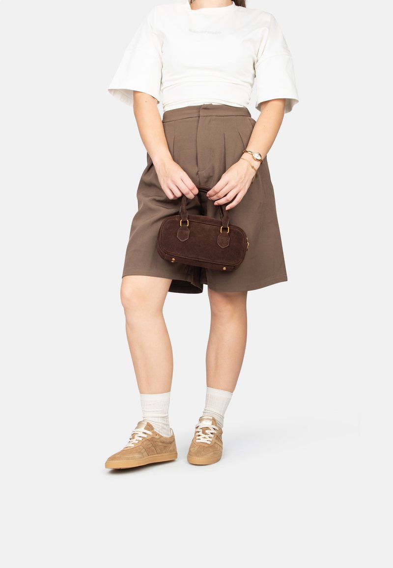 Bolso de ante marrón con asas cortas, combinado con una blusa blanca de manga corta y una falda marrón de talle alto. Zapatillas ligeras y calcetas hasta la altura del tobillo.