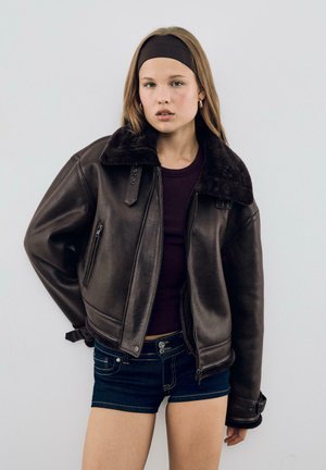 Hnedá kožená bomber bunda s umelou ovčou kožušinou na golieri, so zipsom a patentným zapínaním, spárovaná s tmavými džínsovými šortkami.