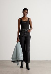 Débardeur noir avec logo, jeans noirs évasés taille haute, tenant une doudoune bleu clair. Des bottes à talons larges noires complètent le look.