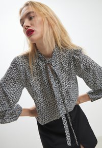 Blusa a righe bianco e nero con collo annodato, maniche lunghe a sbuffo e un'apertura a chiave. Presenta un tessuto liscio e una vestibilità comoda.