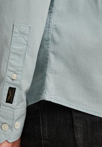 Camisa de botones azul claro con botones blancos, dobladillo angular y etiqueta negra. Presenta costuras visibles y un acabado de algodón texturizado.