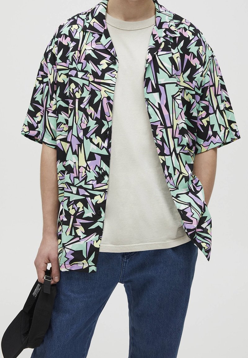 Personne portant une chemise à manches courtes à motif géométrique pastel par-dessus un t-shirt blanc et un jean bleu, tenant une casquette noire dans une main.
