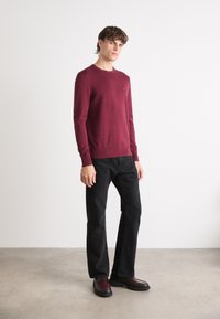 Burgunderfarget strikkegenser med lange ermer og ribbestrikkede mansjetter, kombinert med svarte flagrende jeans og svarte slip-on sko med maroon-detaljer.