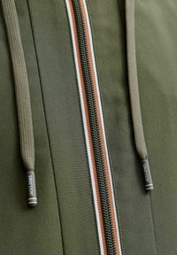 Maslinasto zelena jakna s prednjim patentnim zatvaračem, bijelim i narančastim prugama, teksturiranim vezicama i brendiranim metalnim detaljima.