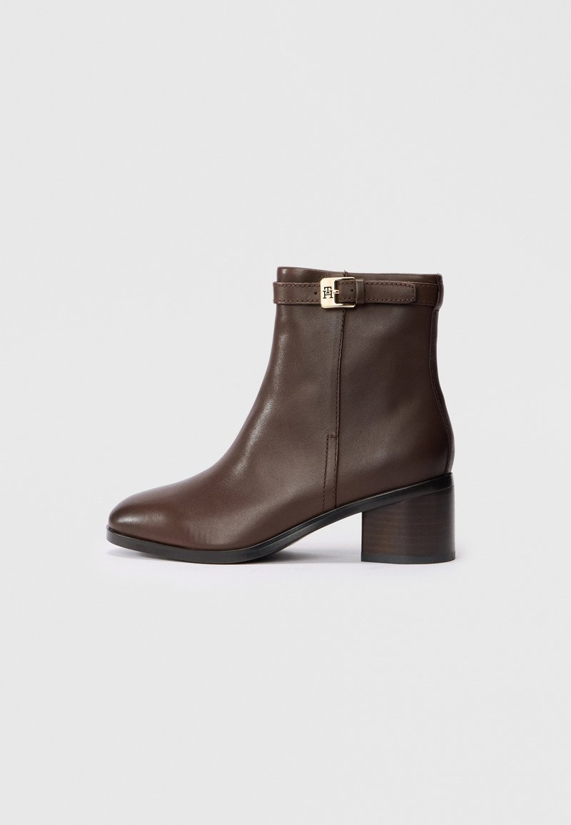 Bottine en cuir marron avec talon bloc et bride à boucle dorée sur le côté, sur un fond gris clair uni.