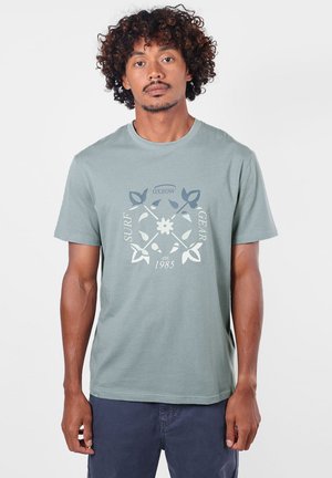 Homme aux cheveux bouclés portant un t-shirt vert clair avec un logo d’équipement de surf et un pantalon bleu foncé, debout devant un fond uni.