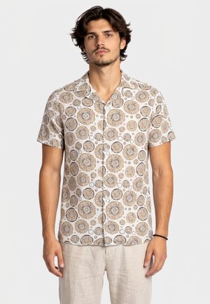 Uomo che indossa una camicia a maniche corte con bottoni e motivi circolari beige e pantaloni chiari su uno sfondo semplice.