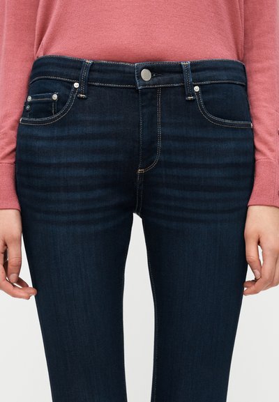 Σκούρο μπλε τζιν με κλασικό σχεδιασμό πέντε τσεπών, ασημένια μεταλλικά στοιχεία και ραμμένες λεπτομέρειες κατά μήκος των ραφών.