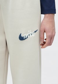 Nike világosszürke anyagú melegítőnadrág, navy kék hímzett swoosh logóval és "nike" felirattal. Simább textúra oldalsó zsebekkel.