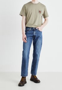 Olivgrön T-shirt med en rävgrafik på bröstet, tillsammans med blå raka jeans och bruna snörade stövlar.