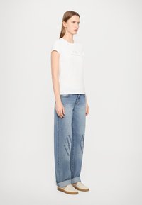 T-shirt bianca in cotone con logo, abbinata a jeans in denim azzurro chiaro oversize con una stampa sottile sulle gambe e scarpe beige.