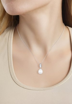 Femme portant un collier en chaîne argentée avec un pendentif en forme de goutte de perle blanche sur un haut beige à col rond.