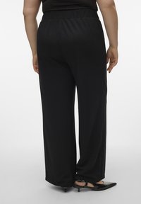 Pantalons larges noirs avec une taille élastique, présentant une texture lisse. Portés avec des talons aiguilles noirs, mettant en valeur un design épuré et minimaliste.