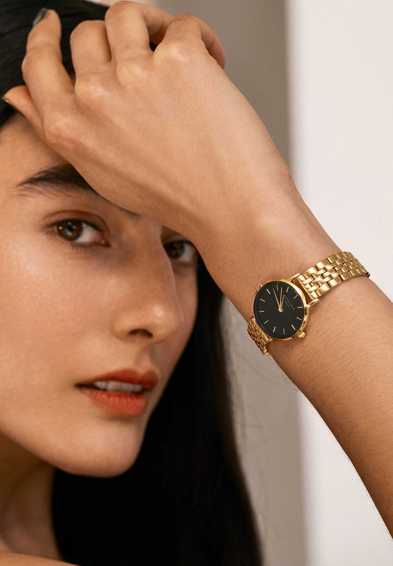 Orologio in metallo dorato con quadrante nero, dotato di un bracciale a rete e indicatori delle ore minimalisti, indossato su un polso su uno sfondo neutro.