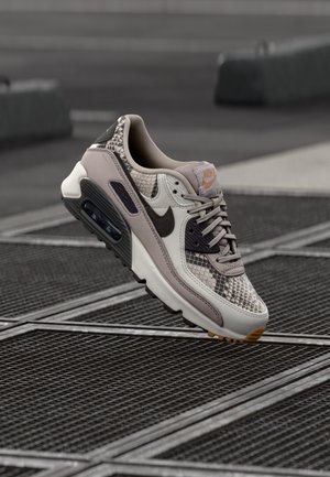Pojedynczy beżowo-brązowy sneaker Nike ze wzorem skóry węża i czarnym swooshem, unoszący się nad metalową kratą na miejskiej ulicy.