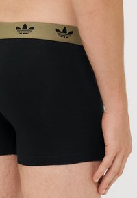 Črne bombažne bokserice z širokim olivno zelenim pasom, ki ima črno dvignjeno logotip in črtasto zasnovo, gladko teksturo in oprijet kroj.