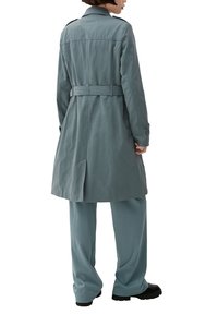 Długi, pasek w talii płaszcz trench w miękkim, matowym materiale, w kolorze niebiesko-skalnym, z tylną wentylacją i zapięciami na guzikach przy rękawach. W zestawie z dopasowanymi spodniami.