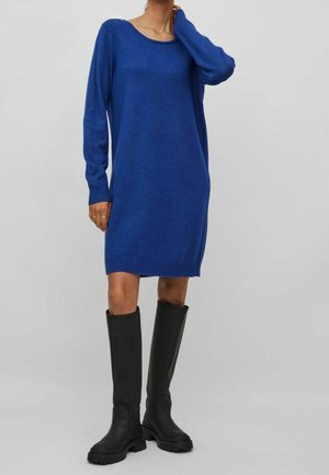 Kobieta ubrana w niebieską, dzianinową sukienkę o długości do kolan z długim rękawem, w połączeniu z czarnymi kozakami za kolano, stojąca z jedną ręką dotykającą szyi.