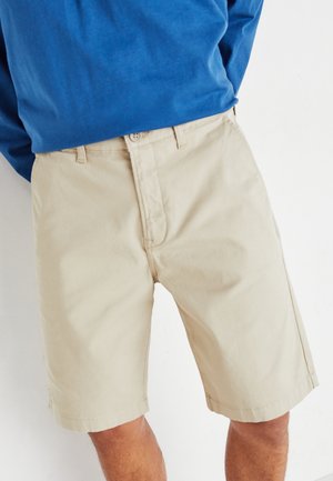 Shorts en coton beige avec une coupe plate, des poches latérales et des ourlets cousus, associés à une chemise bleu à manches longues.