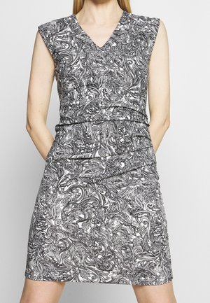 Robe sans manches à motif cachemire noir et blanc avec col en V et taille froncée portée par une personne les mains dans les poches.