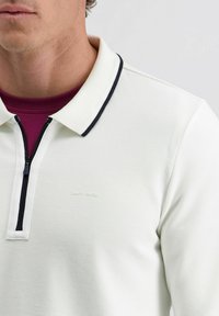 Man in een witte gestructureerde poloshirt met zwarte afwerking en rits, over een paarse ronde hals trui, voor een effen achtergrond.