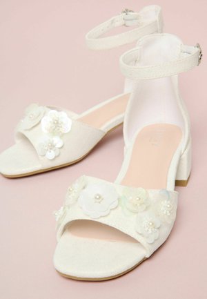 Fehér nyitott orrú blokksarkú cipők bokapántokkal, fehér virágapplikációkkal és gyöngyös díszítéssel rózsaszín háttéren.